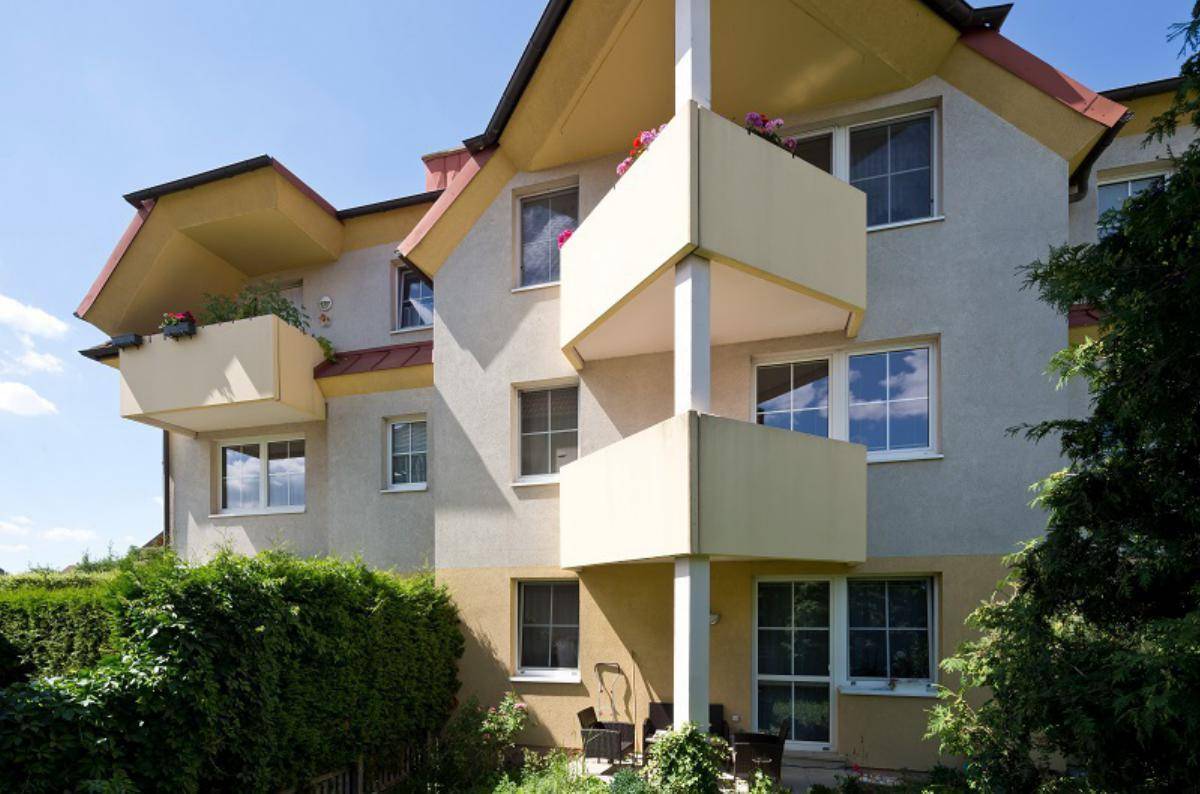 Fassade eines Mehrfamilienhauses mit mehreren Balkonen und großen Fenstern, umgeben von Grün.