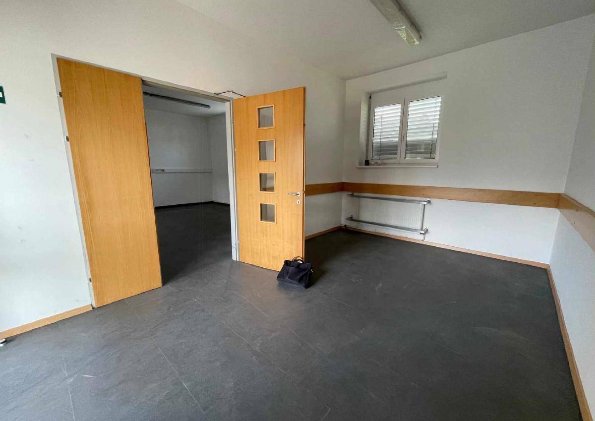 Leerer Raum mit dunklem Boden, weißen Wänden und einer Holztür, die zu einem weiteren Bereich führt.