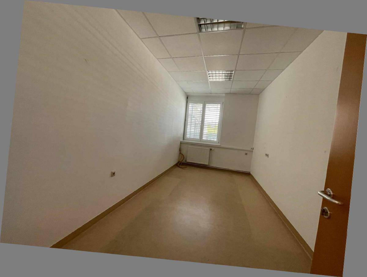 Langer, schmaler Büroraum mit hellem Laminatboden, weißen Wänden und einem Fenster mit Jalousien.