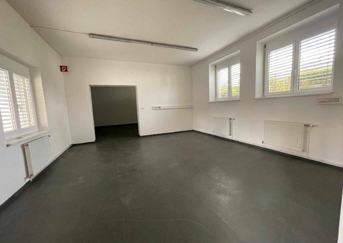 Geräumiger, leerer Gewerberaum mit weißen Wänden, dunklem Boden und großen Fenstern mit Jalousien.