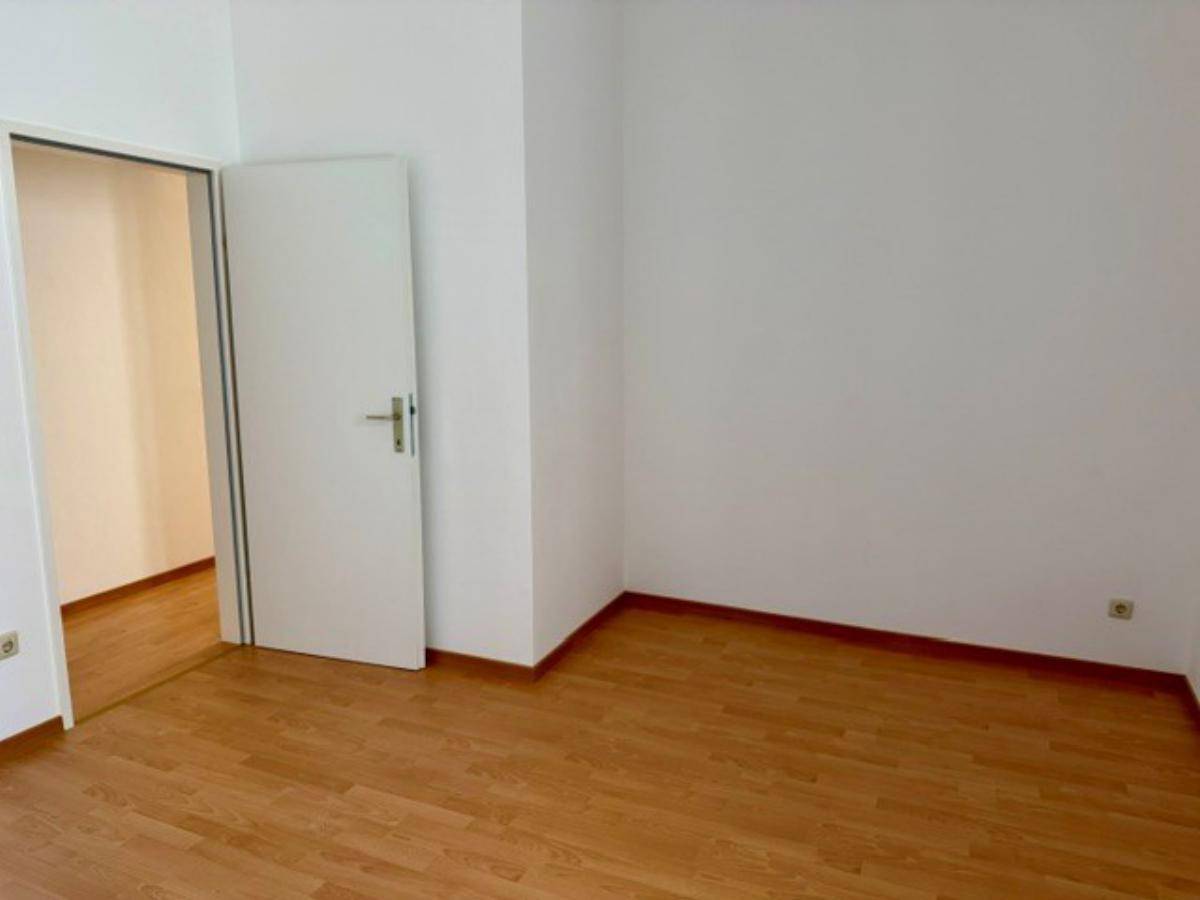 Leeres Zimmer mit Laminatboden, weißen Wänden und einer offenen Tür zu einem weiteren Raum.