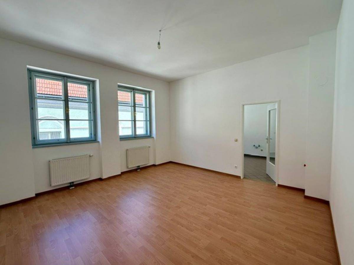 Helles, leeres Zimmer mit Laminatboden, weißen Wänden und zwei großen Fenstern.