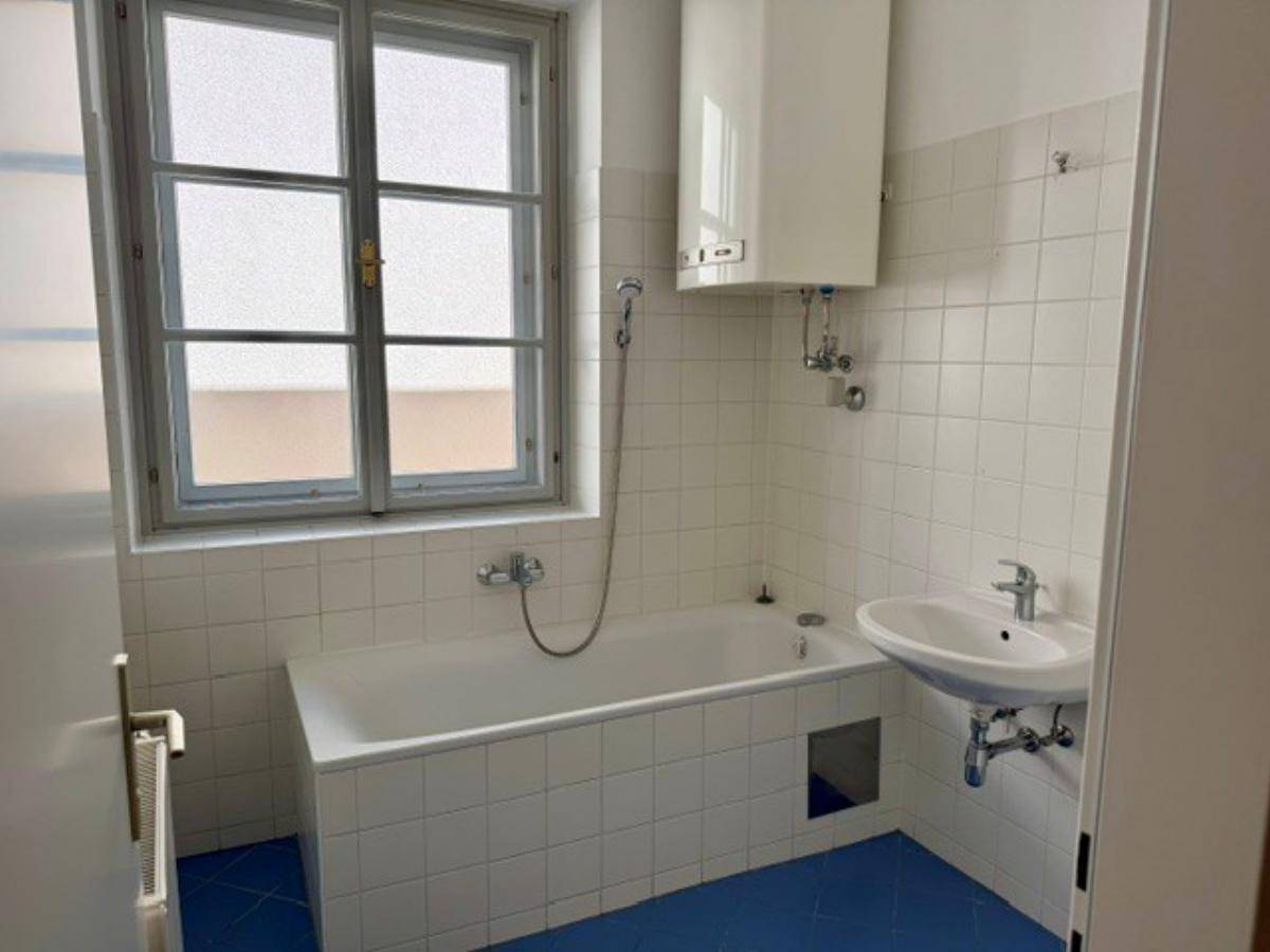 Einfaches Badezimmer mit Badewanne, Waschbecken, Boiler und einem Fenster.