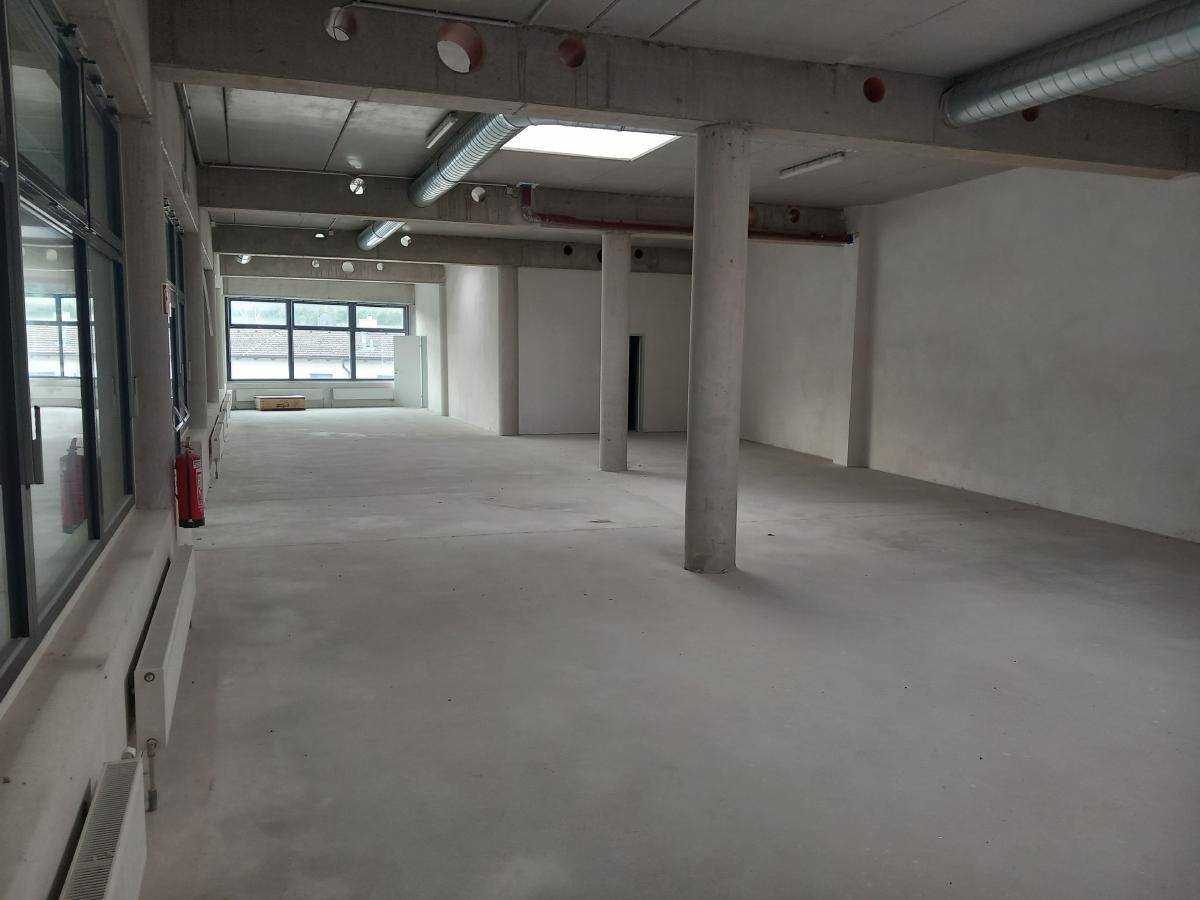 Großer, leerer Gewerberaum mit Betonwänden, hohen Decken und großen Fenstern, die viel Tageslicht hereinlassen.