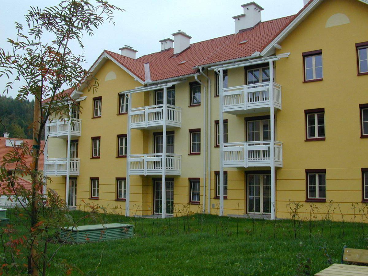 Neubau-Mehrfamilienhaus mit gelber Fassade, roten Dächern und Balkonen, umgeben von Grünflächen.
