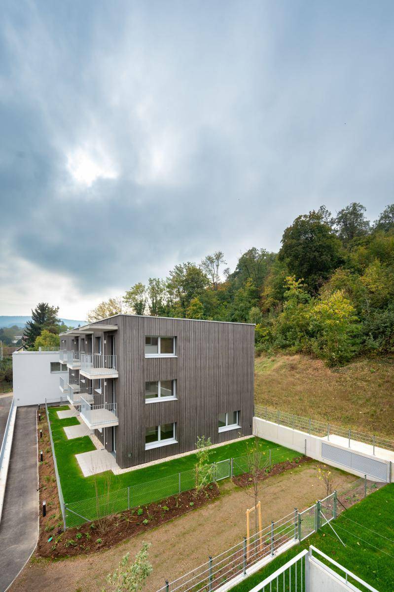 Neubau-Wohngebäude mit dunkler Holzverkleidung und Balkonen, eingebettet in eine grüne Hügellandschaft.
