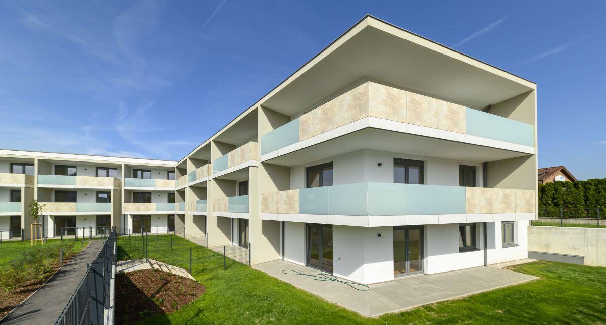 Eckansicht eines modernen Wohngebäudes mit mehreren Balkonen und ebenerdigen Terrassen, umgeben von Rasen.