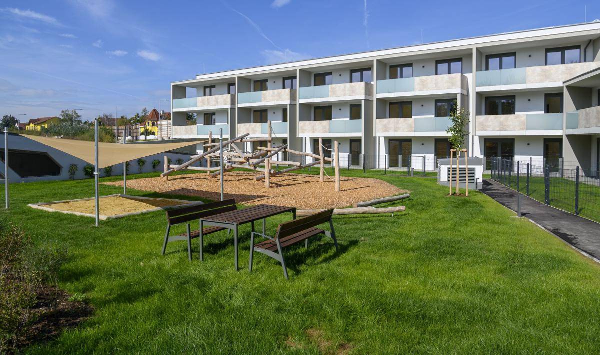 Moderne Wohnanlage mit Spielplatz und Sitzgelegenheiten im Freien, umgeben von gepflegten Grünflächen.