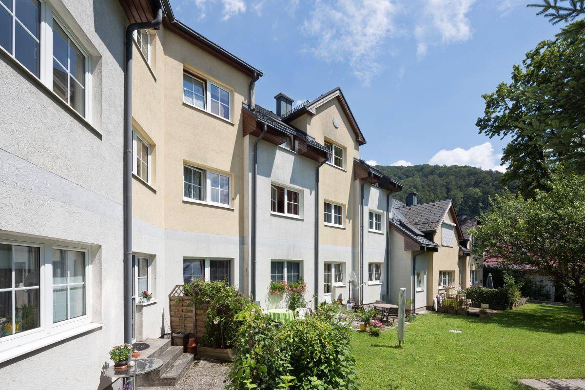 Seitenansicht eines Wohnhauses mit kleiner Terrasse, Rasenfläche und Blick auf bewaldete Hügel.