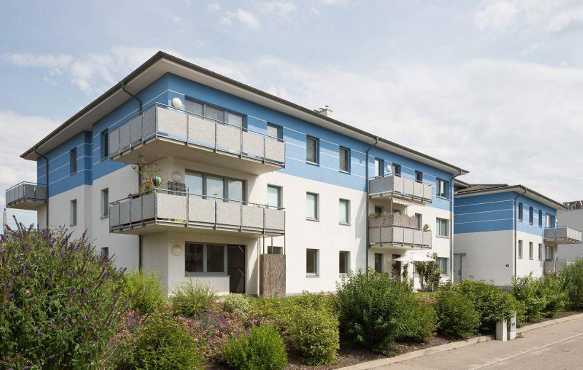 Ansicht eines modernen Apartmentkomplexes mit blauen Akzenten, Balkonen und üppiger Bepflanzung im Vordergrund.