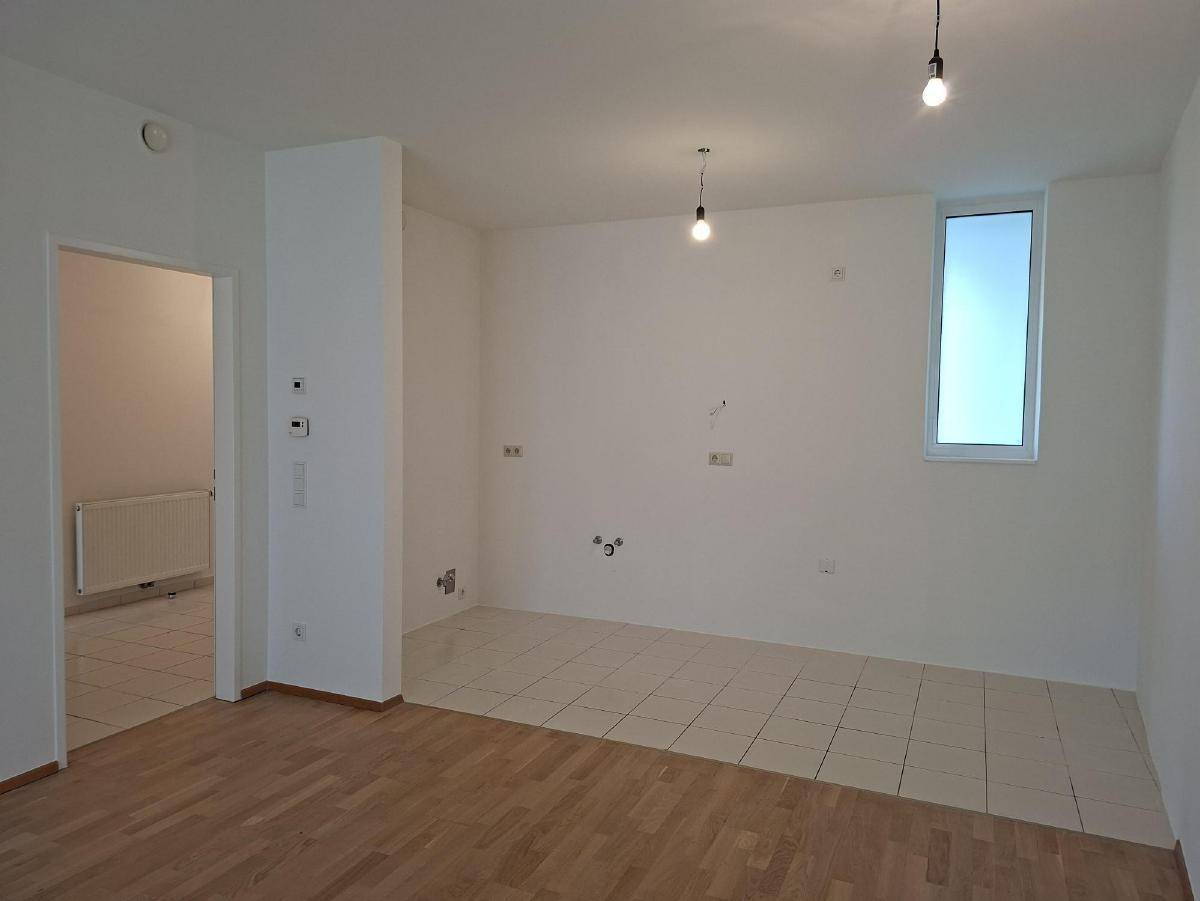 Offener Wohnbereich mit Holzboden und Fliesen, vorbereitet für eine Küche, mit einem hohen Fenster.