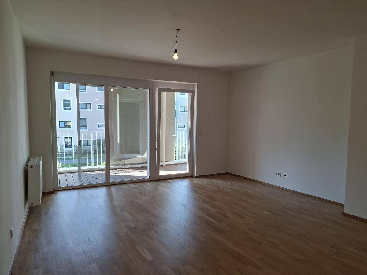 Helles, leeres Wohnzimmer mit Holzboden und großem Schiebefenster zum Balkon.