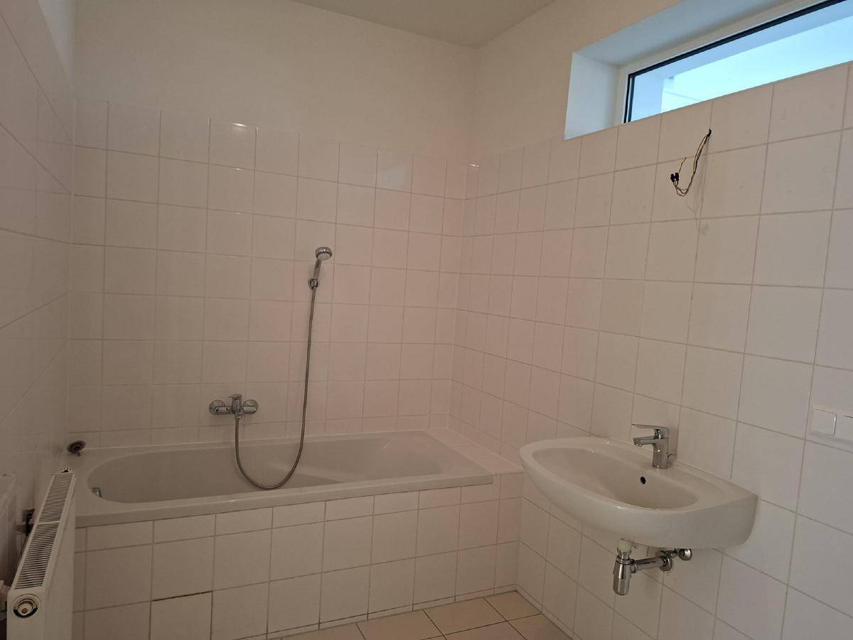Badezimmer mit weiß gefliesten Wänden, Badewanne mit Duschfunktion und einem Waschbecken unter dem Fenster.