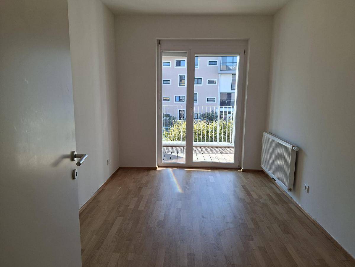 Helles Schlafzimmer mit Holzboden, Heizkörper und Zugang zu einem Balkon durch eine Glastür.