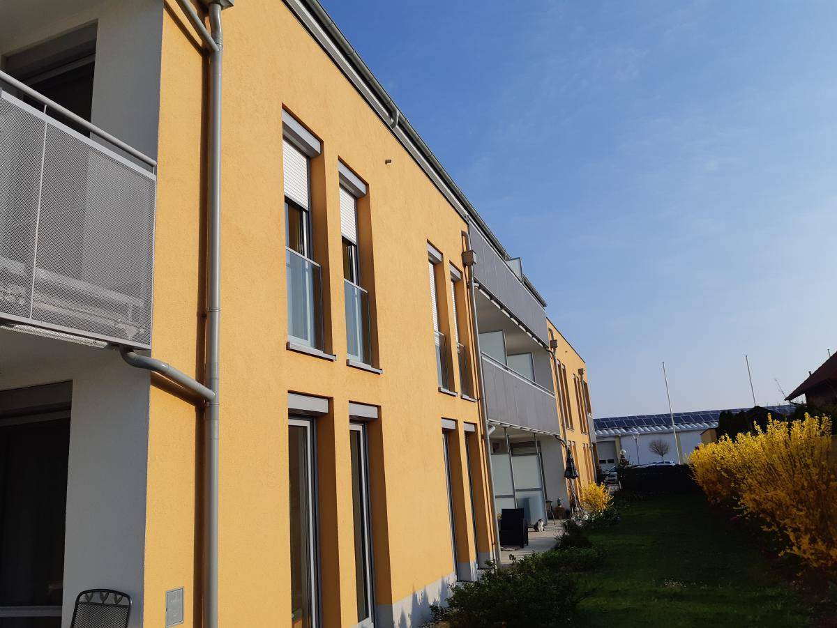 Sonnige Fassade eines modernen Wohnhauses mit Balkonen und grüner Bepflanzung.