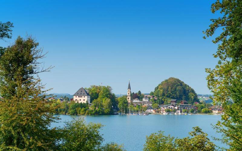 Malerische Ansicht einer Stadt am See mit Schloss, Kirchturm und bewaldeten Hügeln, umrahmt von Bäumen.