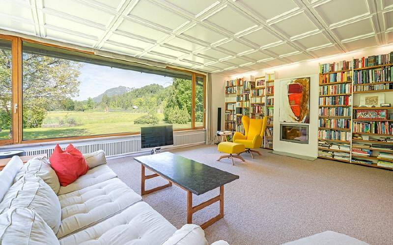 Helles Wohnzimmer mit großer Fensterfront, Blick ins Grüne und umfangreichen Bücherregalen.