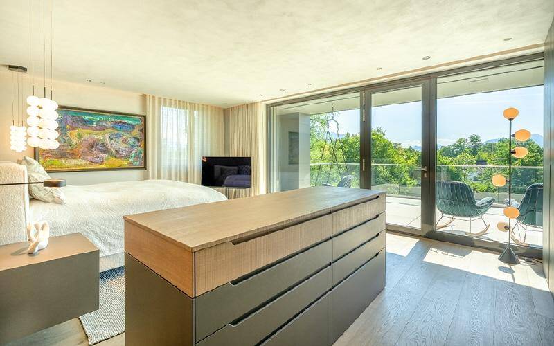 Helles Schlafzimmer mit großem Bett, moderner Kommode und raumhohen Schiebetüren zum Balkon mit Grünblick.