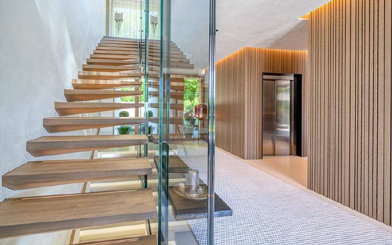 Elegantes Treppenhaus mit Holzstufen und Glasgeländer, daneben ein moderner Aufzug mit Holzverkleidung.