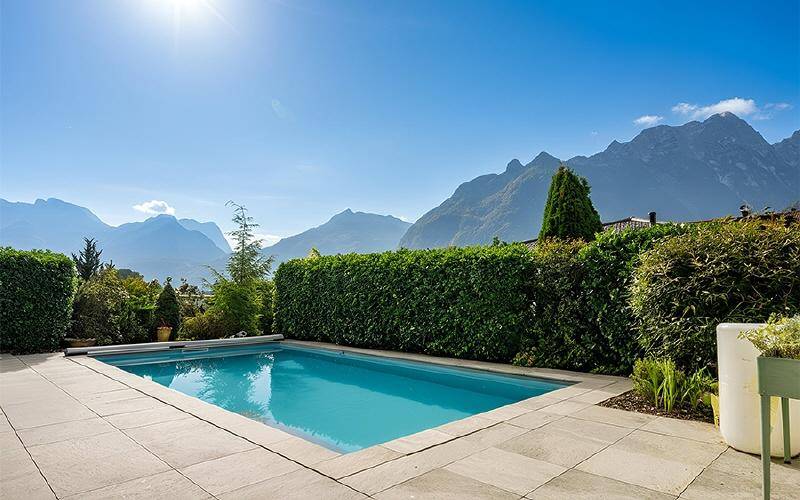 Außenpool mit Bergblick und gepflegter Hecke unter strahlend blauem Himmel.