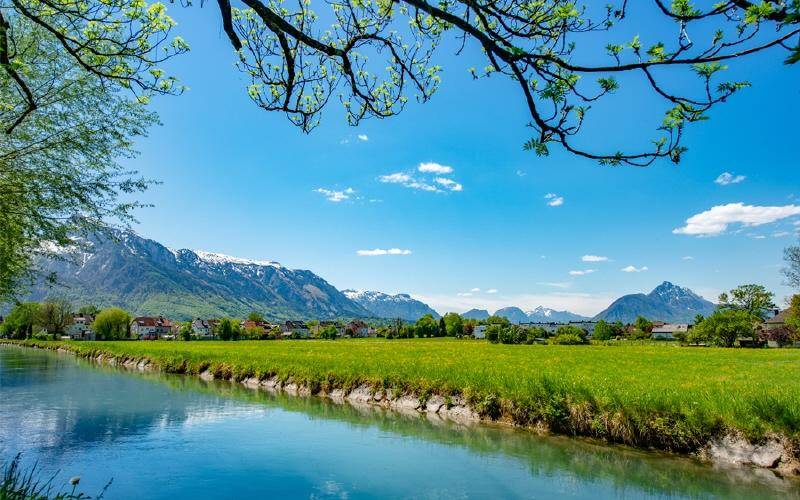 Idyllischer Blick auf einen Fluss, grüne Wiesen und die majestätischen Alpen im Hintergrund.
