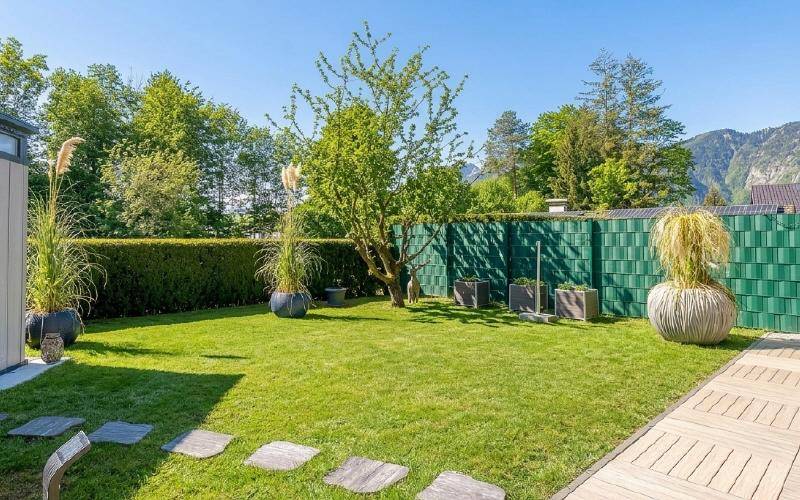 Gepflegter Garten mit Rasenfläche, hohen Zierpflanzen und einem Holzdielenweg unter blauem Himmel.