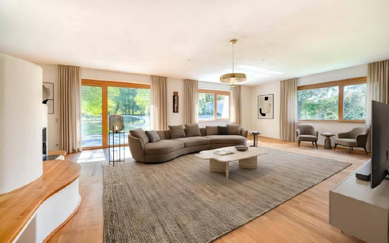 Lichtdurchflutetes Wohnzimmer mit elegantem Sofa, Holzboden und Blick ins Grüne durch große Fenster.