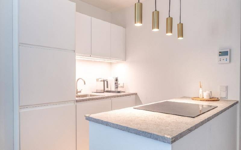 Moderne weiße Küche mit Induktionskochfeld, integrierten Geräten und stilvollen Pendelleuchten.