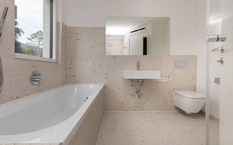 Modernes Badezimmer mit Badewanne, Dusche, wandhängendem WC und beigefarbenen Fliesen.