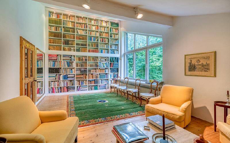 Helles Wohnzimmer mit raumhohem Bücherregal, großen Fenstern und Blick ins Grüne.