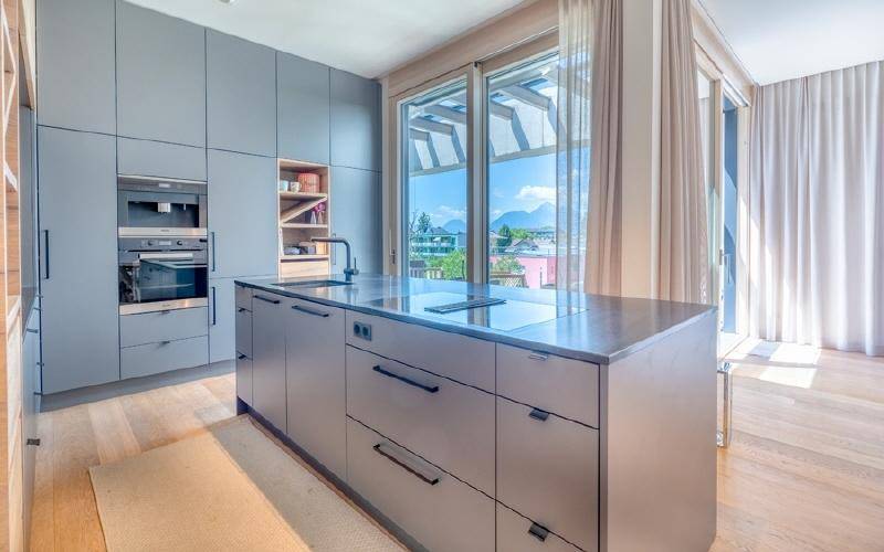 Moderne, graue Einbauküche mit Kochinsel, integrierten Geräten und großem Fenster mit Aussicht.