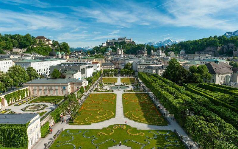 Panoramablick auf eine weitläufige Gartenanlage mit Stadt und schneebedeckten Bergen im Hintergrund.
