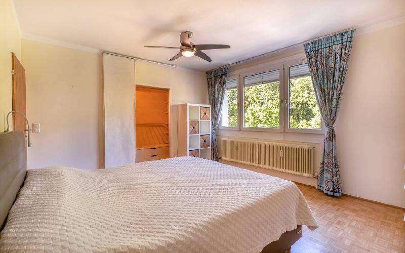 Gemütliches Schlafzimmer mit Doppelbett, Parkettboden, Deckenventilator und großen Fenstern mit Blick ins Grüne.