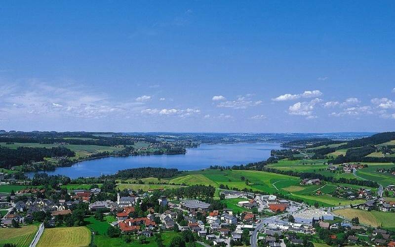 Luftaufnahme eines Sees, umgeben von sanften grünen Hügeln und einer kleinen Stadt im Vordergrund.