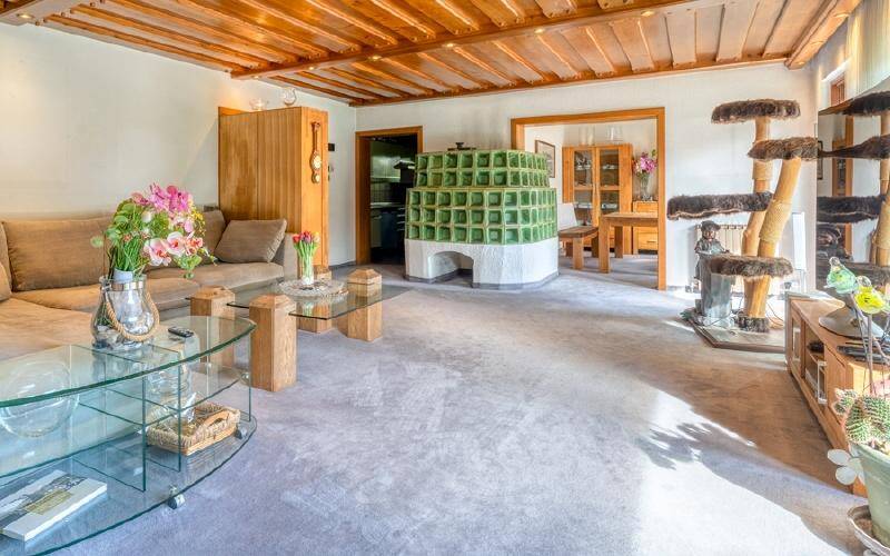Gemütliches Wohnzimmer mit traditionellem Kachelofen, Holzbalkendecke und Teppichboden.