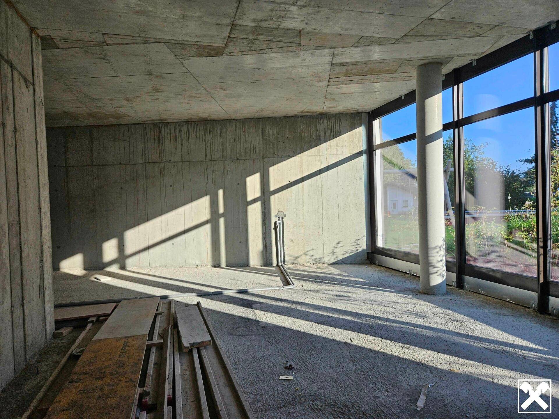Heller, unfertiger Innenraum mit Betonwänden und großen Fenstern, die viel Sonnenlicht hereinlassen.