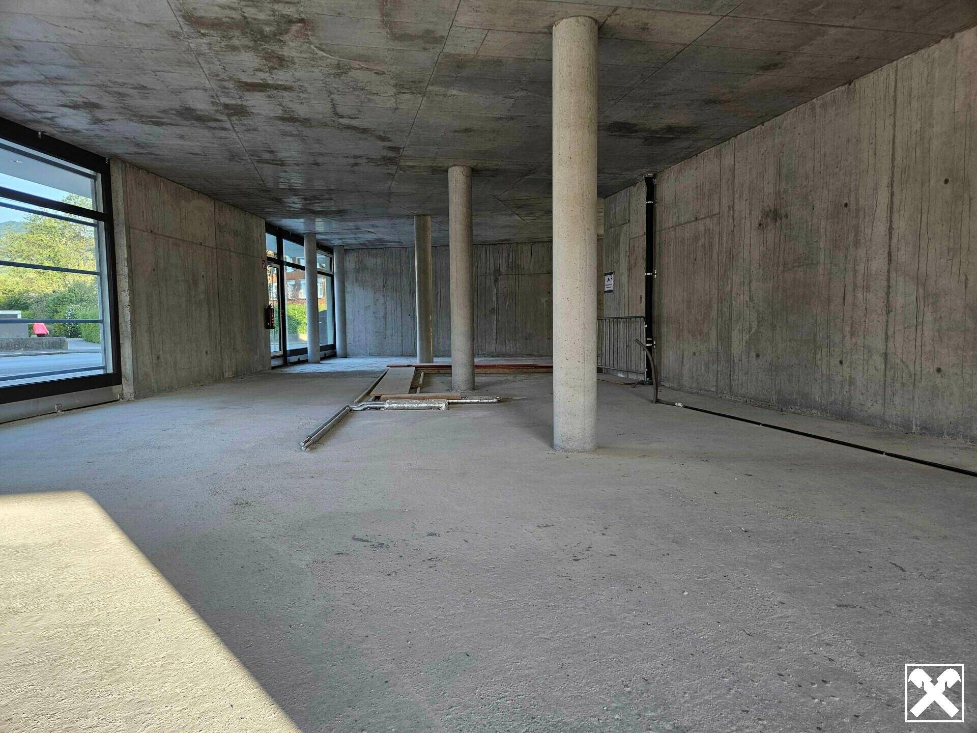 Weiter, unfertiger Raum mit Betonwänden und -boden, gekennzeichnet durch tragende Säulen und viel Tageslicht.