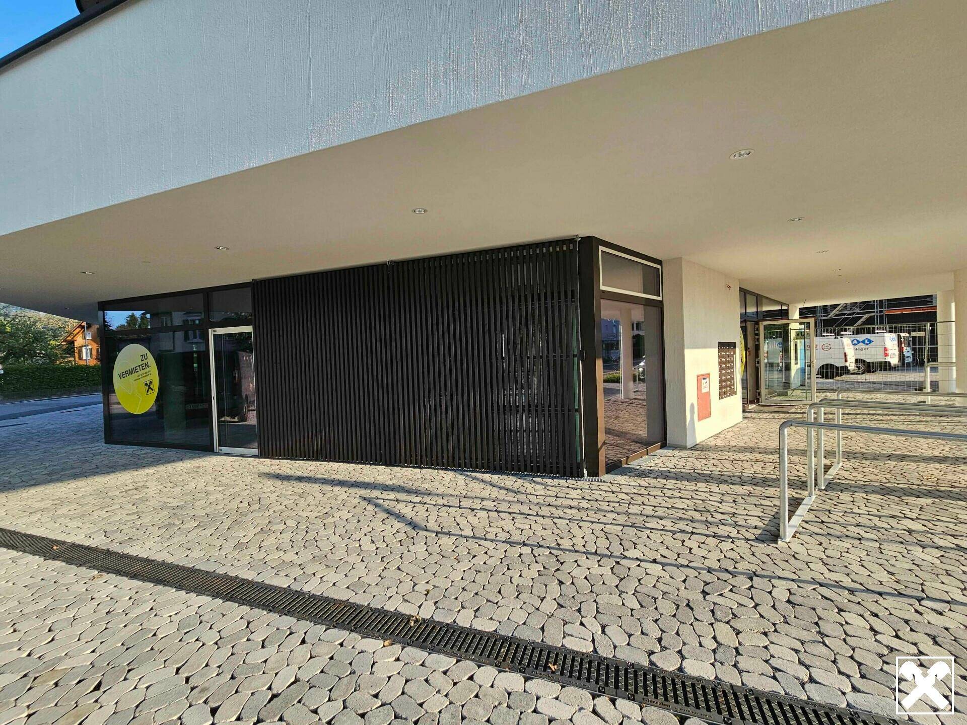 Moderne Geschäftsfassade mit dunklen Holzlamellen und großen Schaufenstern, umgeben von Pflastersteinen.