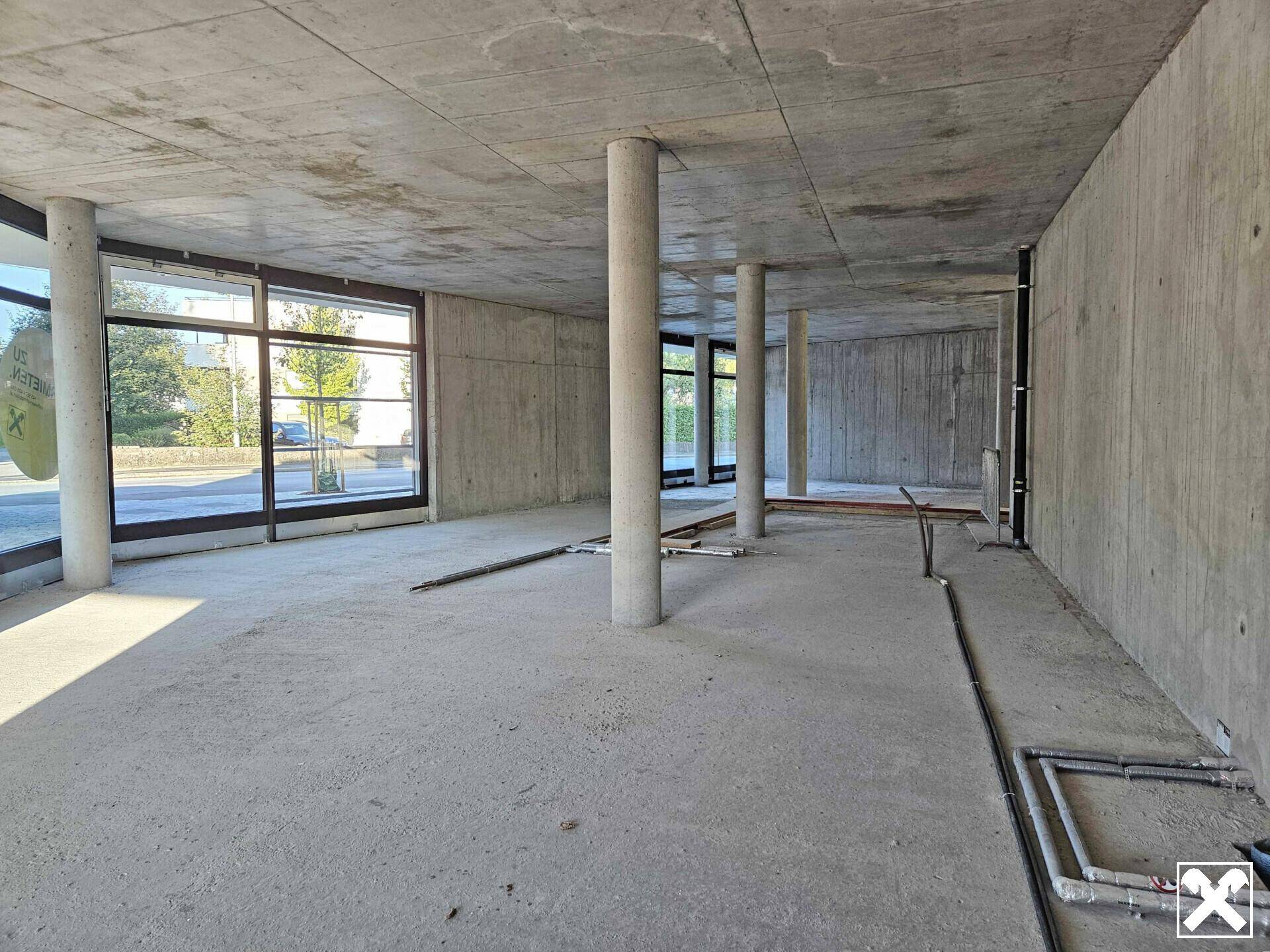 Großer, unfertiger Innenraum mit Betonwänden und -boden, ausgestattet mit Säulen und großen Fenstern.