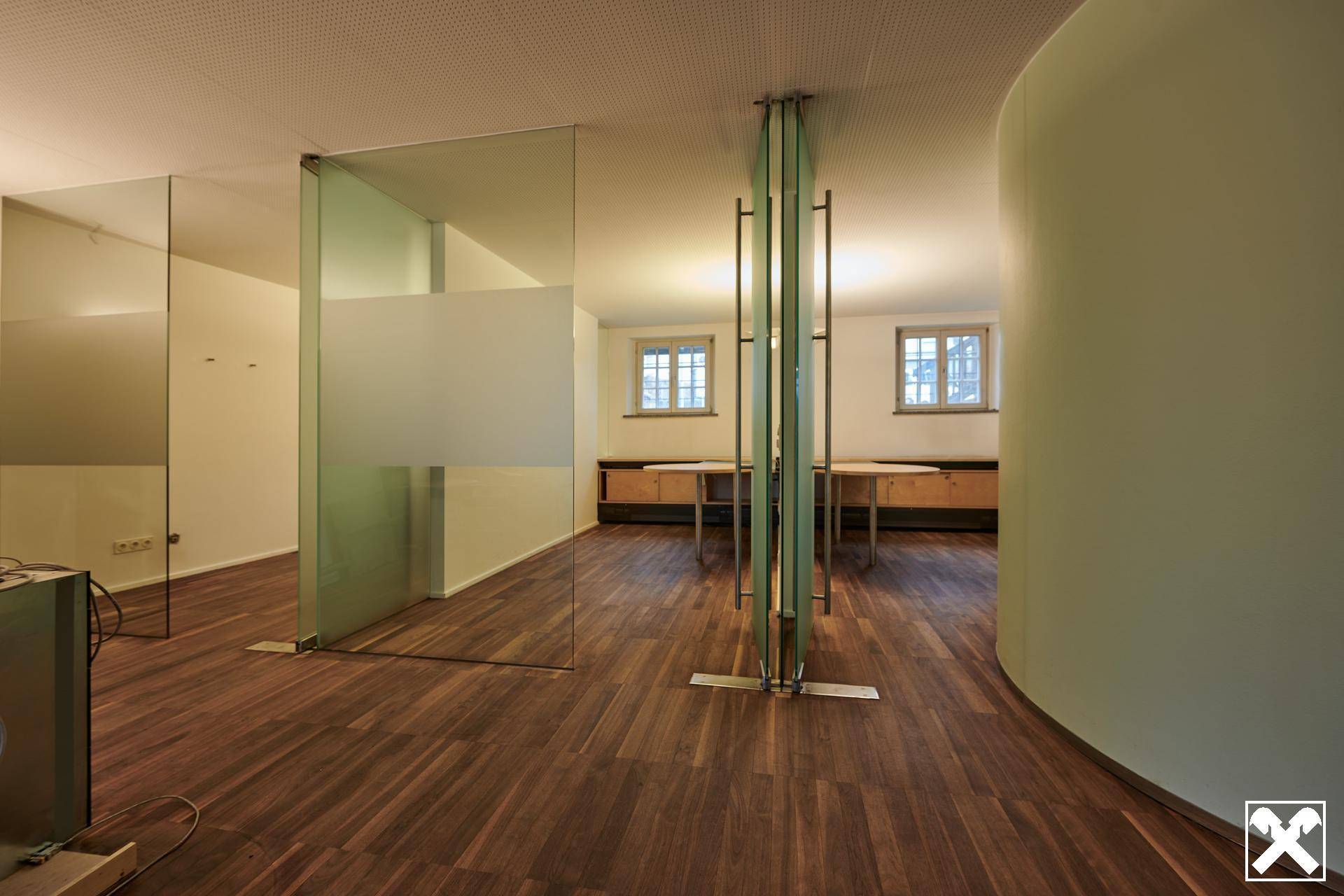 Moderner Büroraum mit Holzboden, Glaswänden als Trennelemente und Fenstern mit Tageslicht.