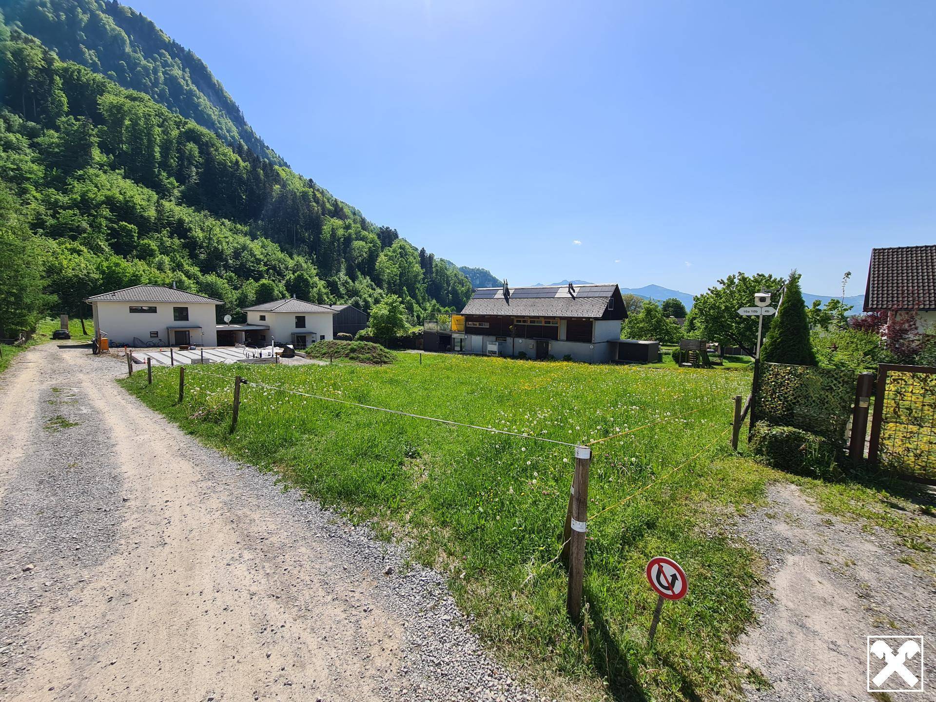 Schotterweg führt zu einer grünen Wiese mit Häusern und beeindruckendem Bergblick.