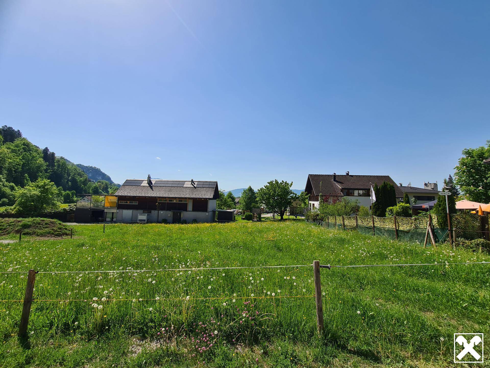 Grüne Wiese mit Blick auf Wohnhäuser und eine bewaldete Bergkette im Hintergrund.