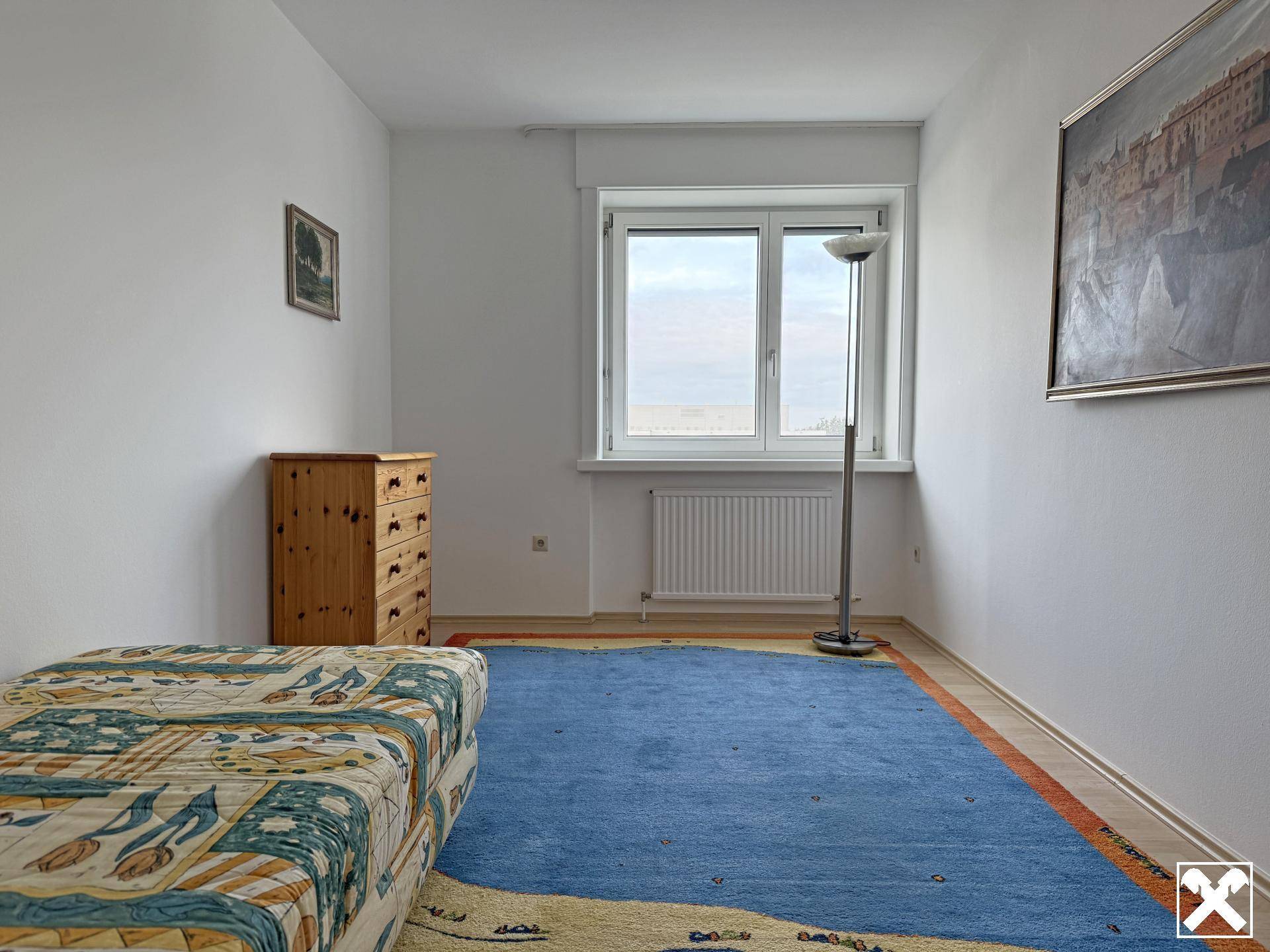 Helles Zimmer mit Fenster, Teppichboden und einfacher Einrichtung.