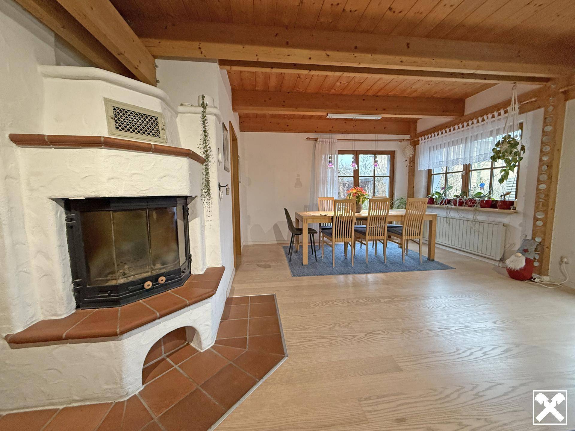 Heller Wohn-Essbereich mit Kamin, Holzbalkendecke und großem Esstisch am Fenster.