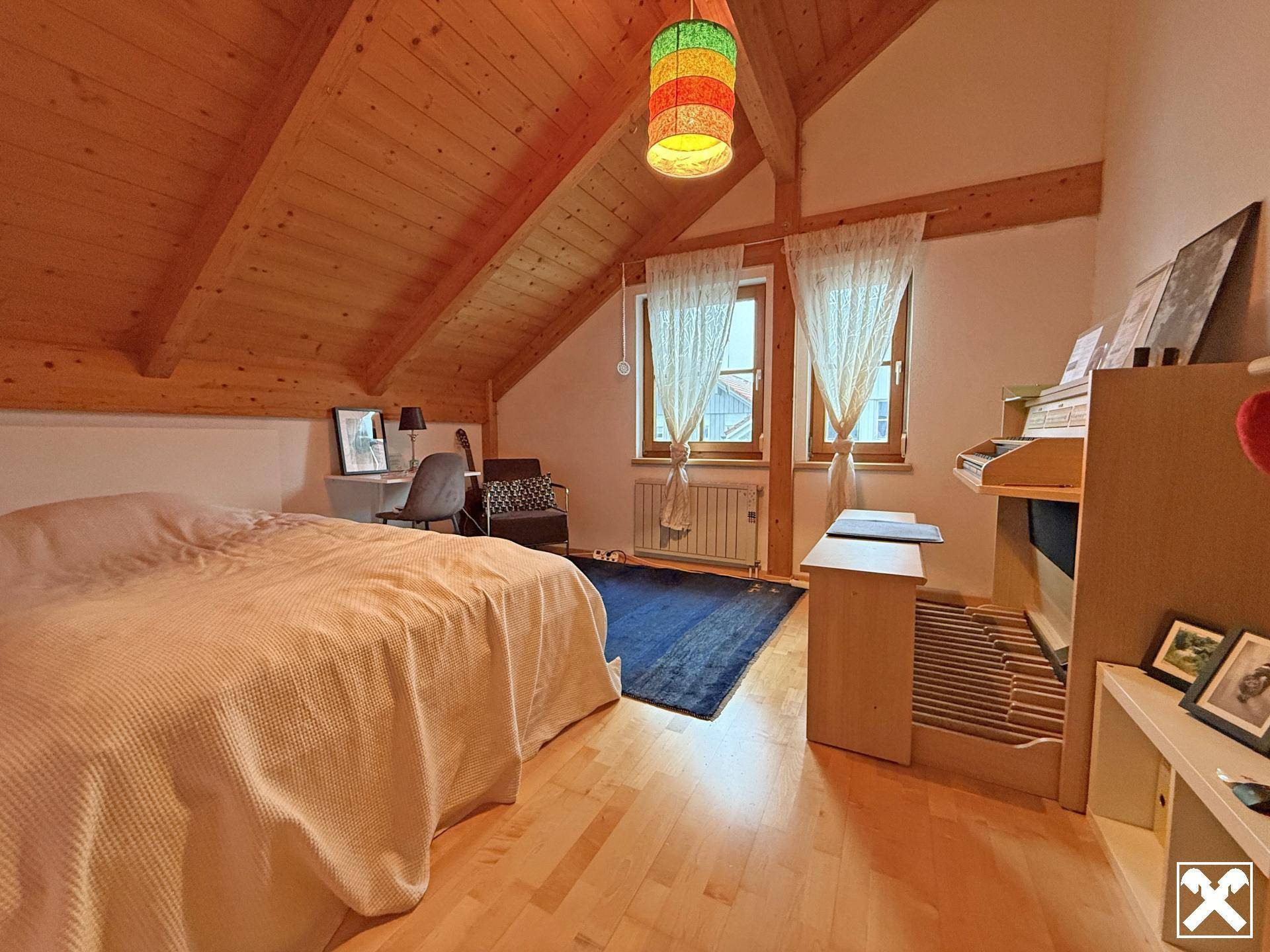 Gemütliches Schlafzimmer im Dachgeschoss mit Holzbalkendecke, Holzboden und zwei Fenstern.