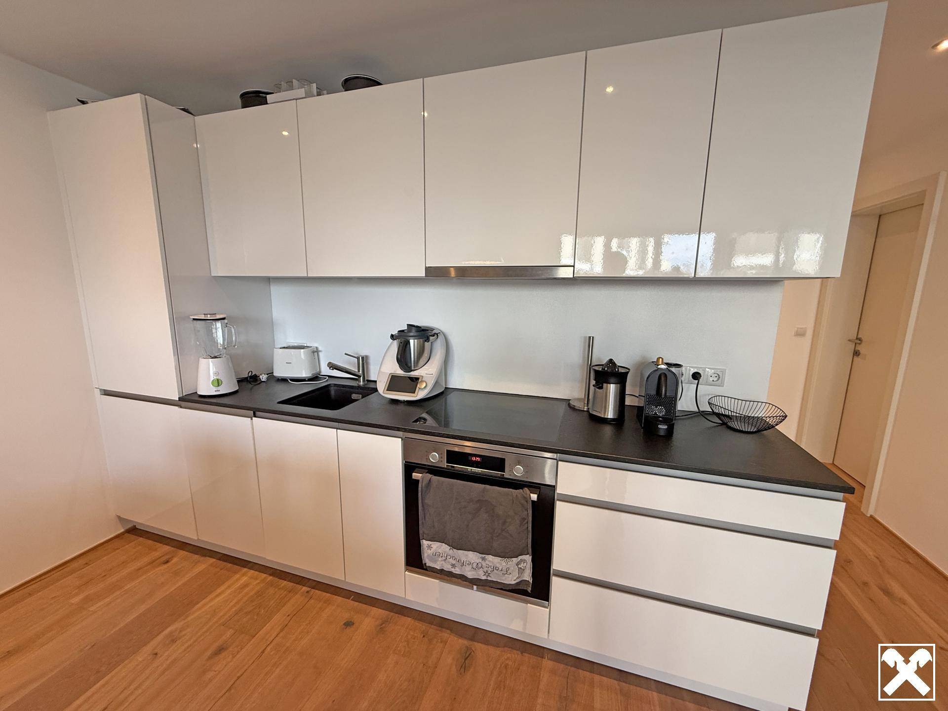 Moderne Küche mit weißen Hochglanzfronten, dunkler Arbeitsplatte und integriertem Backofen und Dunstabzugshaube.