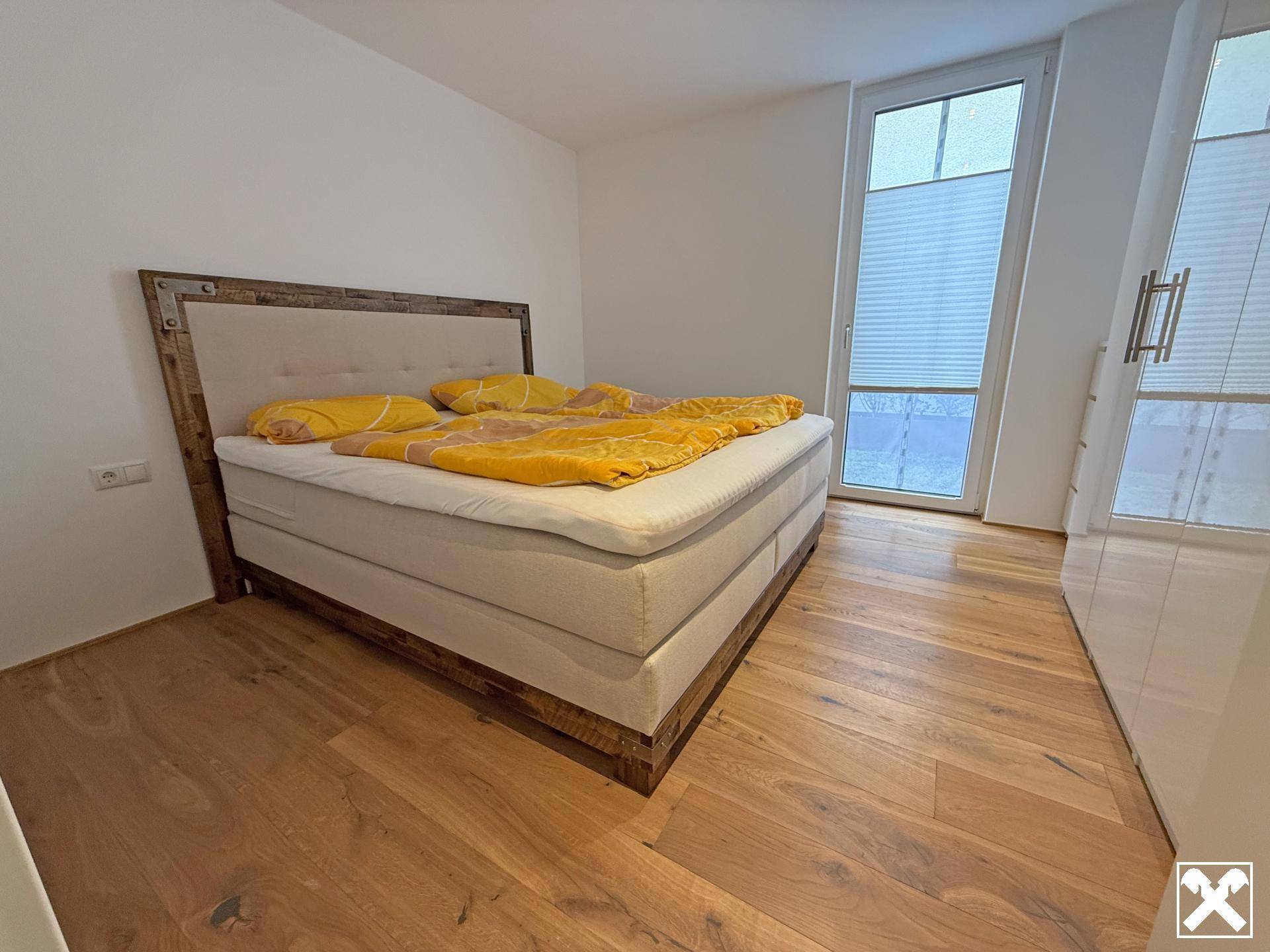Helles Schlafzimmer mit Doppelbett, Kleiderschrank und großem Fenster für viel Tageslicht und Holzboden.