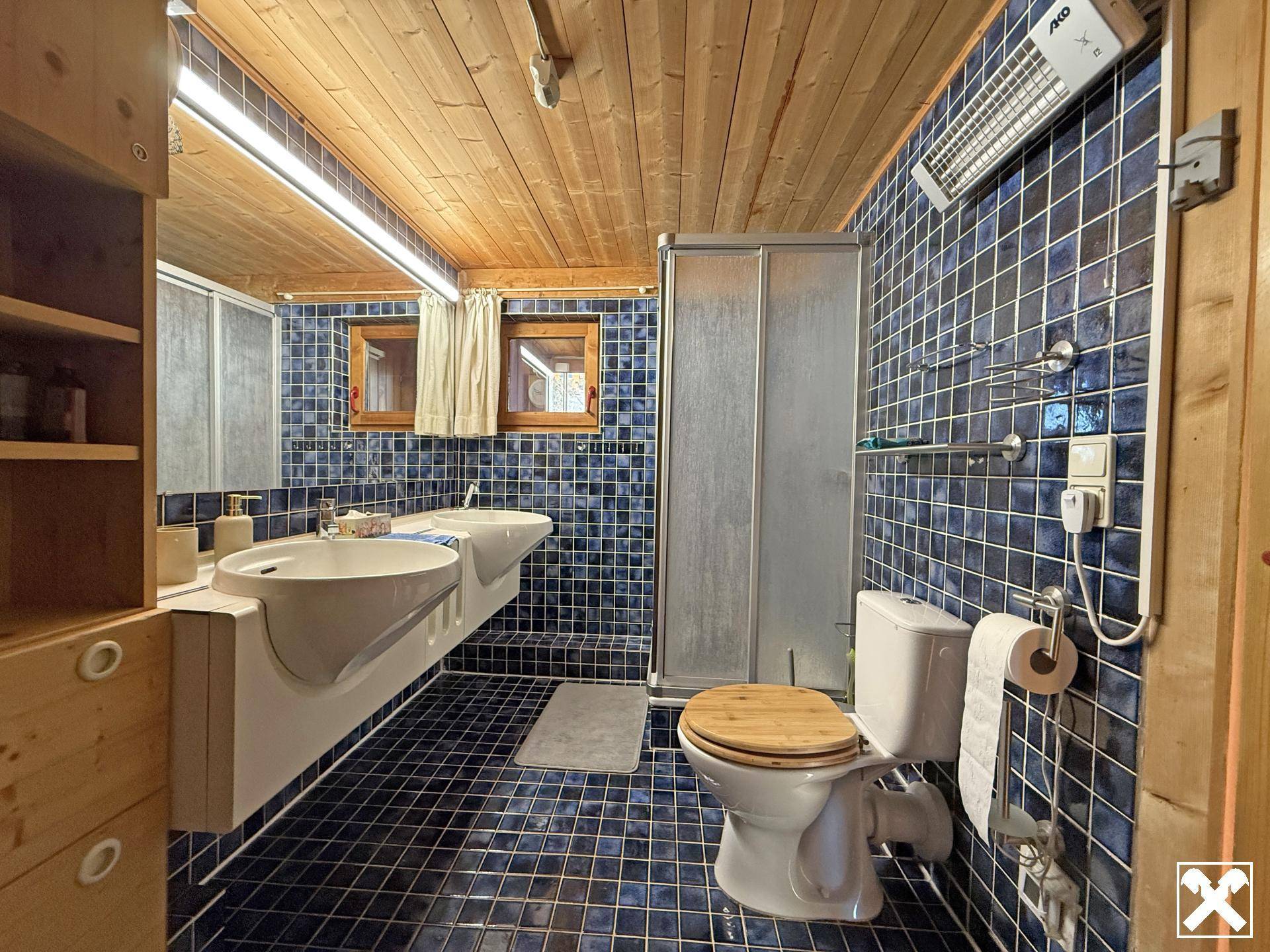 Badezimmer mit blauen Fliesen, Duschkabine, Toilette und Doppelwaschbecken unter einer Holzdecke.