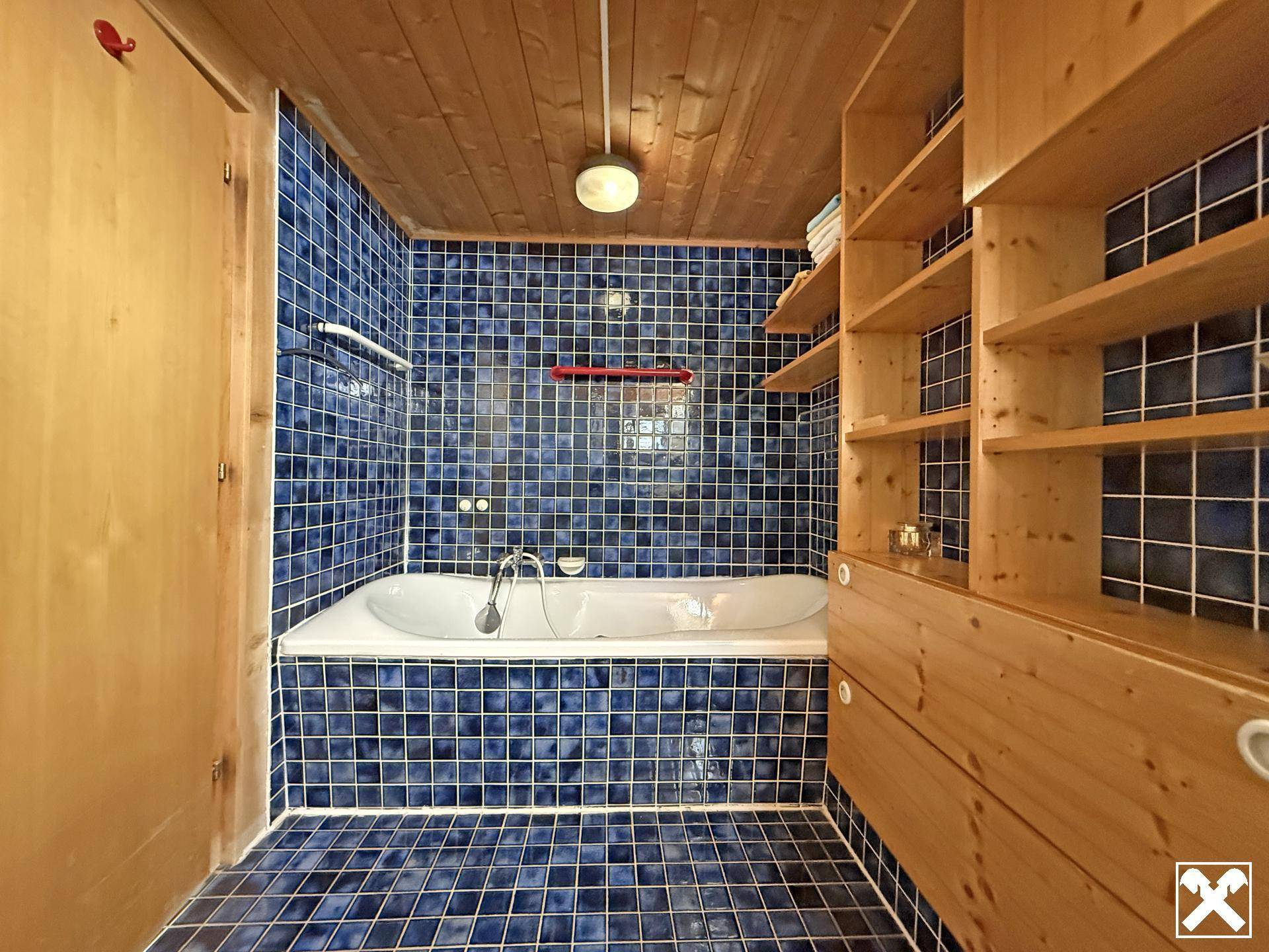 Badezimmer mit blauen Fliesen, Badewanne und praktischen Holzregalen, ergänzt durch eine Holzdecke.