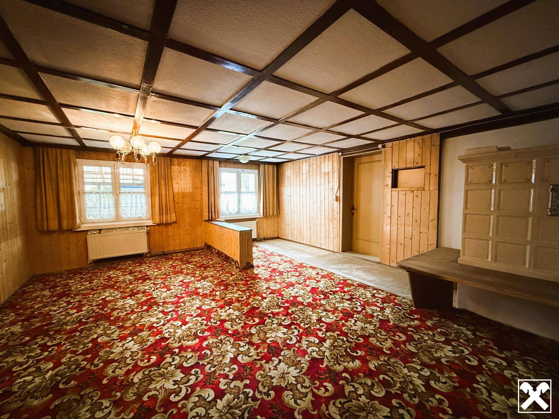 Geräumiges Wohnzimmer mit traditionellem Kachelofen, Holztäfelung und gemusterter Teppichboden.